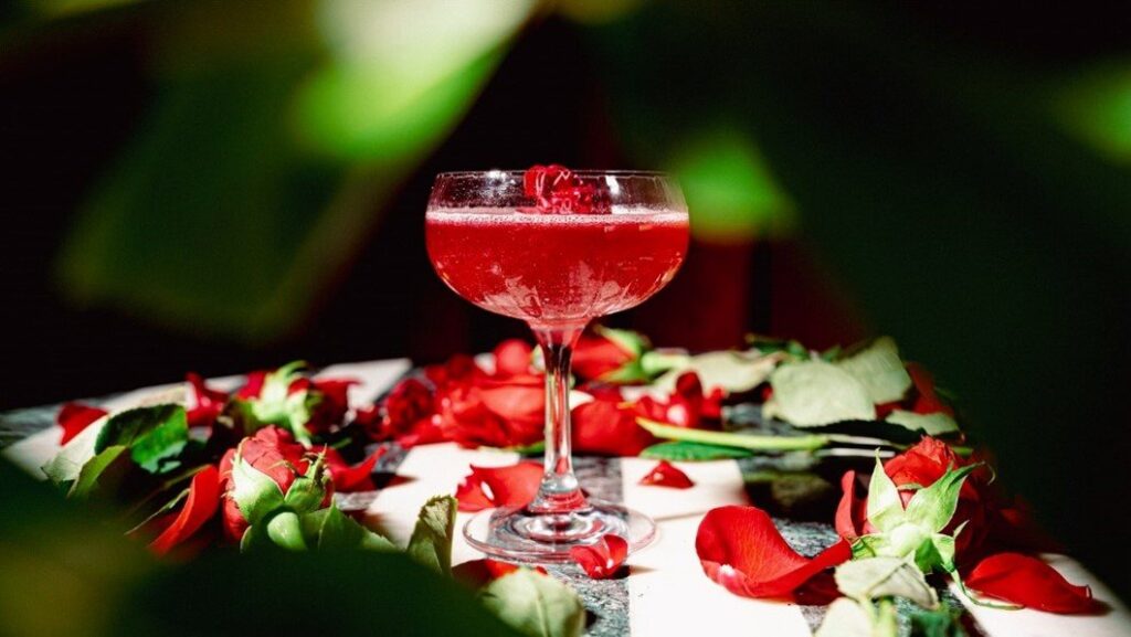 A red martini cocktail in a glass, surrounded by vibrant roses, at Plume in Houston, TX.